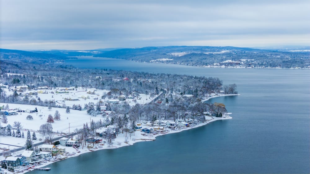 Aerial winter view of snow-covered homes and wooded hills wrapping around the shore of Keuka Lake.