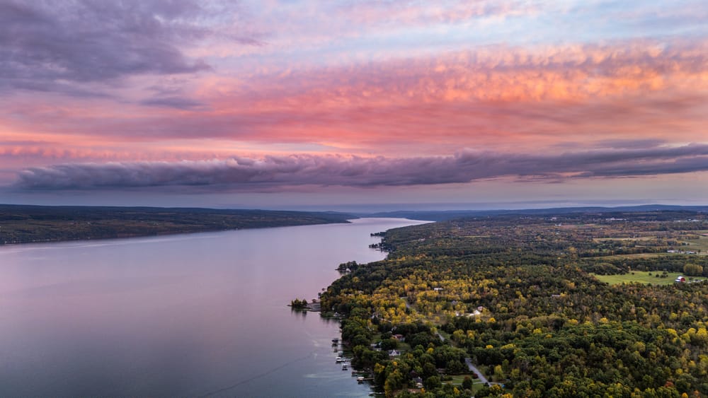 Aerial view down Seneca Lake at sunset with pink clouds, wooded shoreline, and vineyard fields.