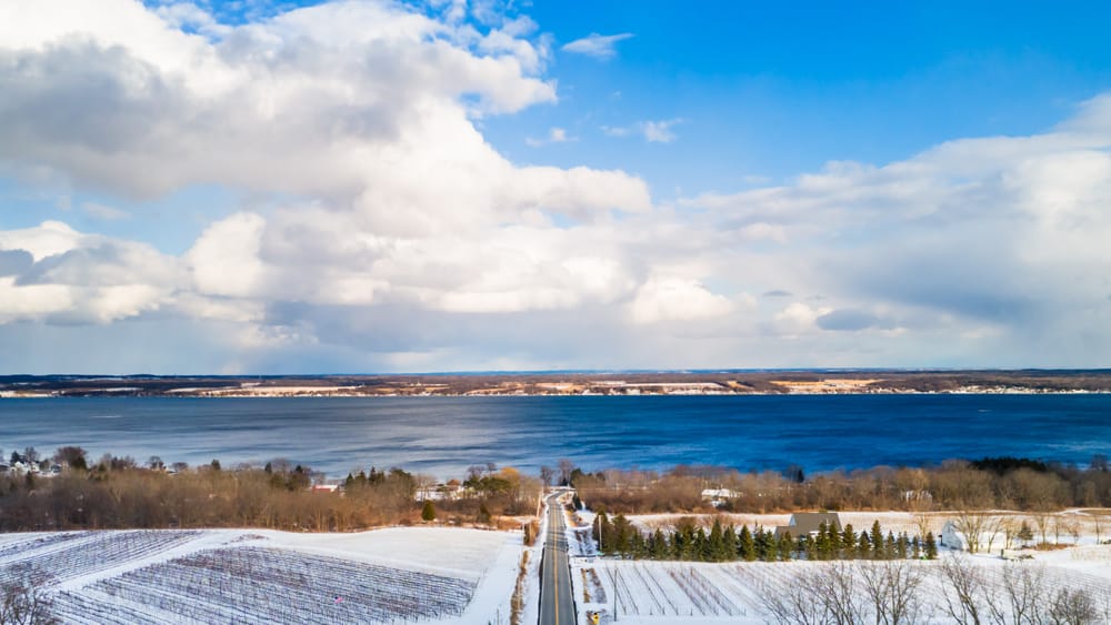Snowy vineyard rows and a straight road descending toward blue Seneca Lake beneath large clouds.