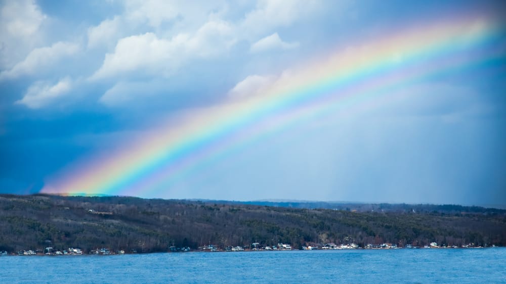 A vivid double rainbow crossing a blue-gray sky above Keuka Lake and its wooded shoreline.