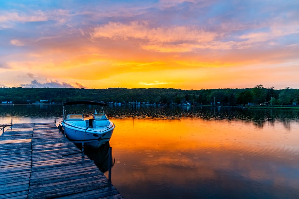 White motorboat tied beside a wooden dock beneath an orange and pink sunset reflected on calm water.